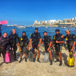 Load image into Gallery viewer, Group of scuba divers posing on a rocky shore in Malta with Bugibba in the background
