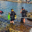 Load image into Gallery viewer, Two scuba divers with equipment standing on rocks by a body of water with buildings in the background.
