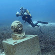 Load image into Gallery viewer, A diver posing behind the divers helmet statue on the reef at Zurrieq, near the Um El Faroud wreck

