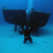 Load image into Gallery viewer, A diver kneeling on the sand in front of the P29 shipwreck on a fun dive in Cirkewwa, Malta
